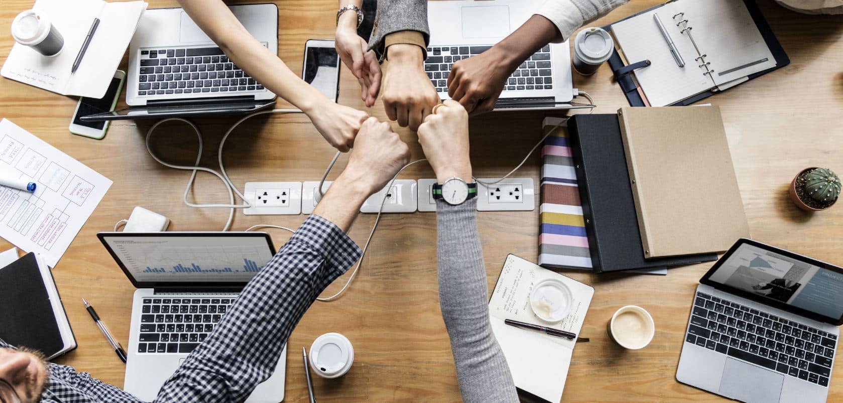 A group of people sitting around a wooden table, each with a laptop in front of them, putting their fists together in the center of the table in a gesture of teamwork